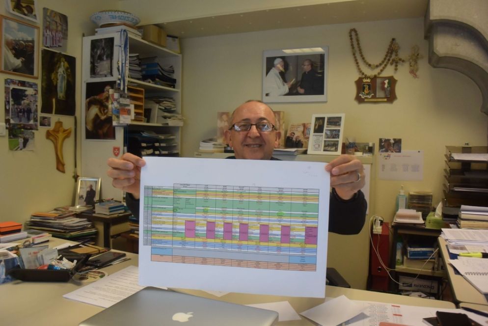 Volto caro a chi segue il Rosario da Lourdes su TV2000, padre Nicola Ventriglia, oblato di Maria Immacolata, cappellanno coordinatore per la lingua italiana, mostra nel suo studio il planning della presenza alla Grotta dei cappellanni del mondo intero