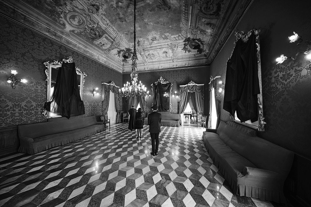 Ragusa, Circolo di Conversazione, 2018. Durante le varie fasi dell’elaborazione del lutto si sospendono le attività quotidiane che normalmente si svolgono tra le pareti domestiche. Anche nel sontuoso “Salone delle Feste” del Circolo di Conversazione a Ragusa, ogni chiacchiera si interrompe e gli specchi vengono coperti per evitare che “imprigionino” l’anima del defunto. Il cromatismo funebre dei lunghi veli neri trionfa dolorosamente sugli arredi e sugli affreschi in stile neoclassico - © Armando Rotoletti