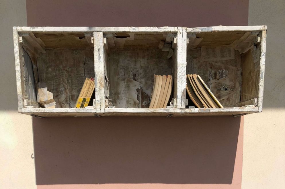 Cassette di legno rivestite di pagine di libro, appese sopra una panchina, sul lungomare di Alassio