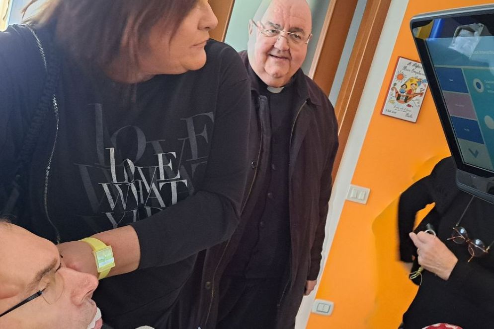 Paola aiuta il marito Stefano con il puntatore oculare che gli permette di comunicare. Con loro, il parroco don Paolo