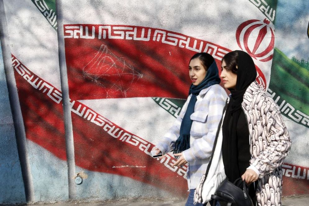 Due ragazze mentre passeggiano di fronte al murale con la bandiera dell’Iran lungo le strade di Teheran - Ansa