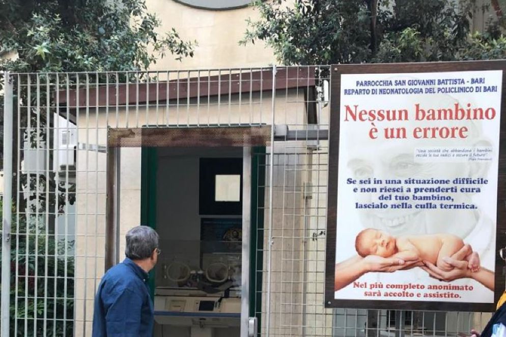 Il parroco davanti alla culla per la vita della parrocchia con il manifesto che pubblicizza lo spazio protetto - Ansa